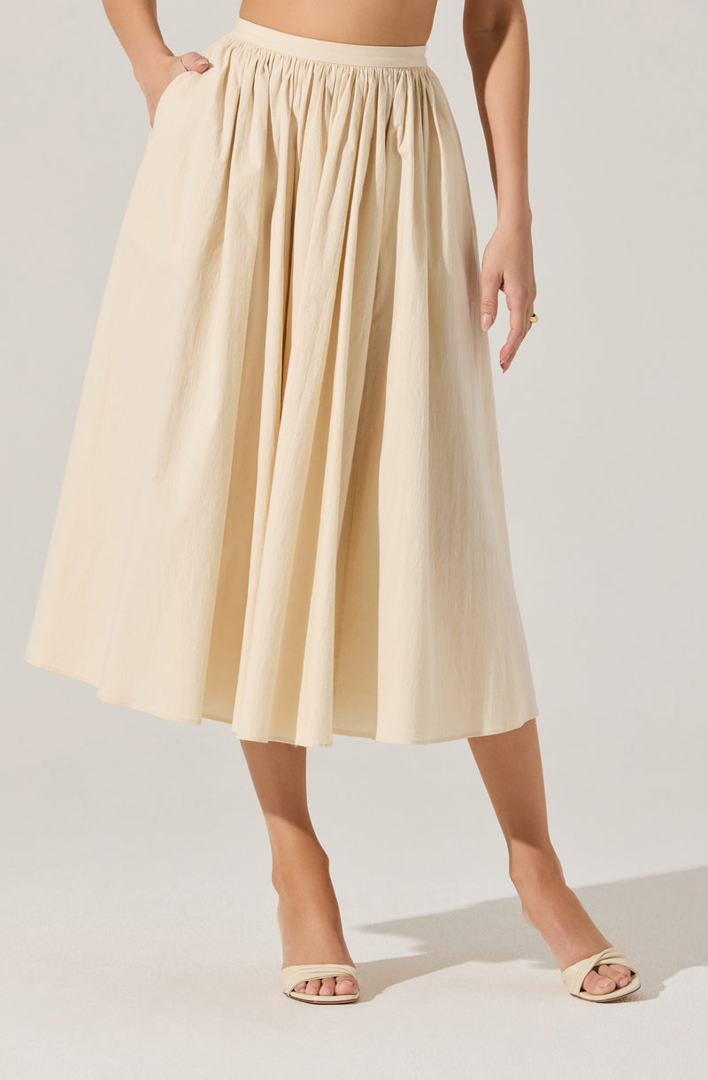 A woman is wearing a high-waisted, full, ankle-length cream-colored skirt with pleats. She pairs the skirt with matching cream-colored open-toe heels.