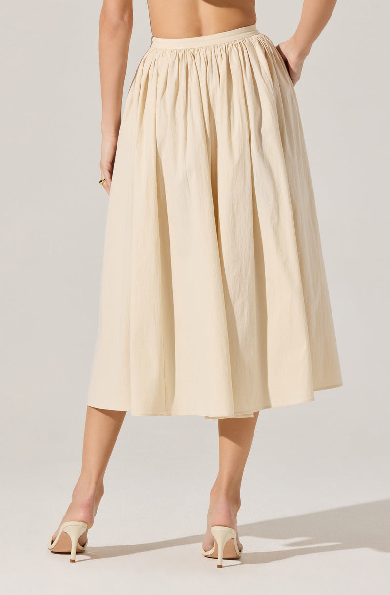 A woman is wearing a cream-colored, ankle-length skirt with a gathered waistband and wide pleats. She pairs the skirt with nude high-heeled shoes. Her hand rests on her hip, showing part of her torso.