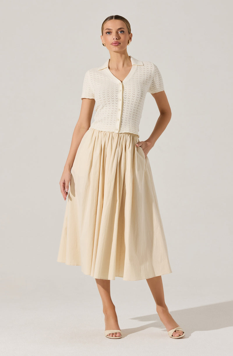 A woman is wearing a short-sleeved, cream-colored knitted cardigan with a V-neck and button closure in the front. The cardigan has a textured, openwork pattern. She pairs it with a high-waisted, long, pleated skirt in a light, neutral color that reaches mid-calf. She completes the outfit with light-colored open-toe heeled sandals.