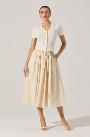 A woman is wearing a short-sleeved, cream-colored knitted cardigan with a V-neck and button closure in the front. The cardigan has a textured, openwork pattern. She pairs it with a high-waisted, long, pleated skirt in a light, neutral color that reaches mid-calf. She completes the outfit with light-colored open-toe heeled sandals.