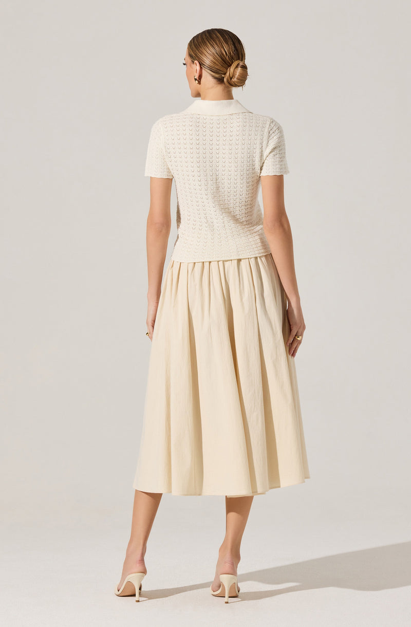 A woman is wearing a short-sleeved, light-colored, textured knit top with a collar. She pairs it with a high-waisted, full-length, beige skirt that has a gathered, pleated design. She is also wearing light-colored high-heeled shoes.