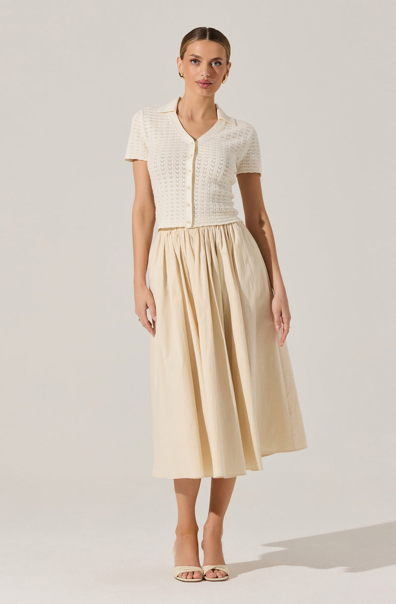 A woman is wearing a short-sleeved, open-knit, cream-colored cardigan with a collar and buttons down the front, paired with a high-waisted, beige, pleated midi skirt. She has on light-colored open-toe heels.