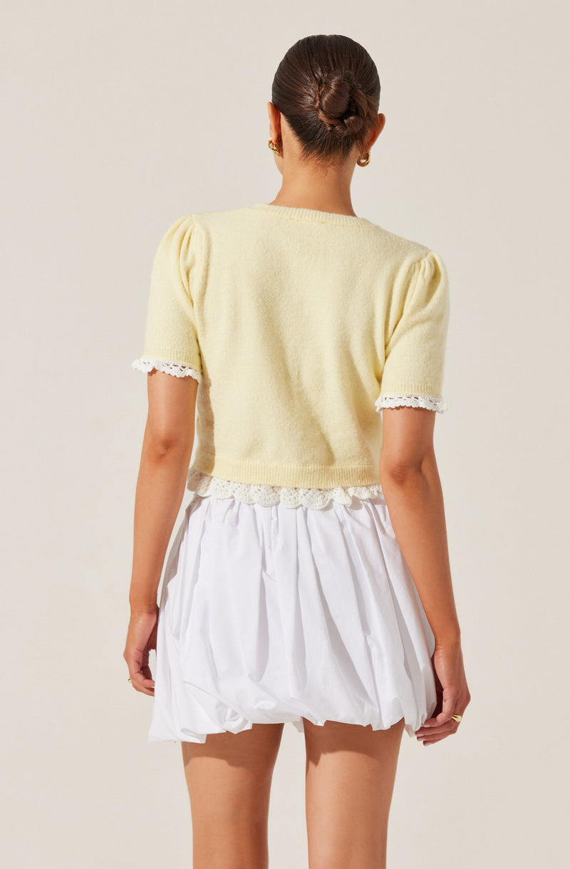 A woman is wearing a short-sleeved, light yellow sweater with a delicate lace trim at the sleeve cuffs and hem. The sweater appears soft and slightly cropped. She pairs it with a white, voluminous, bubble-style skirt that has a gathered, puffy silhouette. Her hair is styled in a neat, twisted bun, and she accessorizes with small, gold earrings.