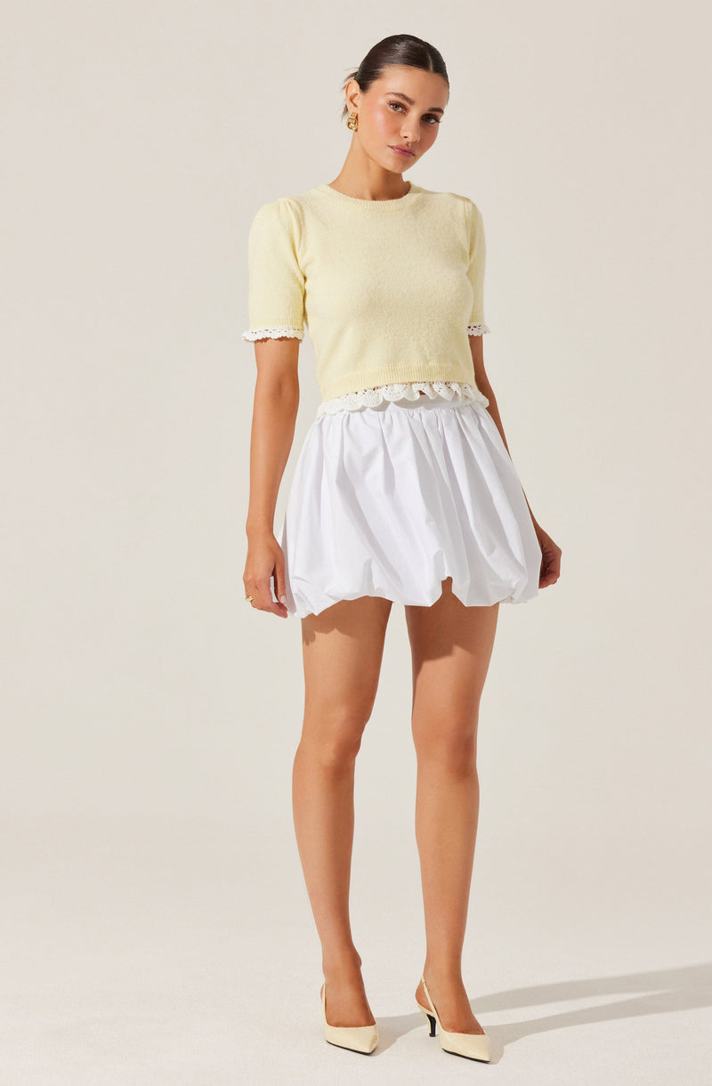 A woman is wearing a light yellow, short-sleeved, cropped knit top with lace trim on the sleeves and hem. She pairs it with a white, puffed, knee-length skirt with an elastic waistband. She completes the outfit with beige high-heeled shoes.