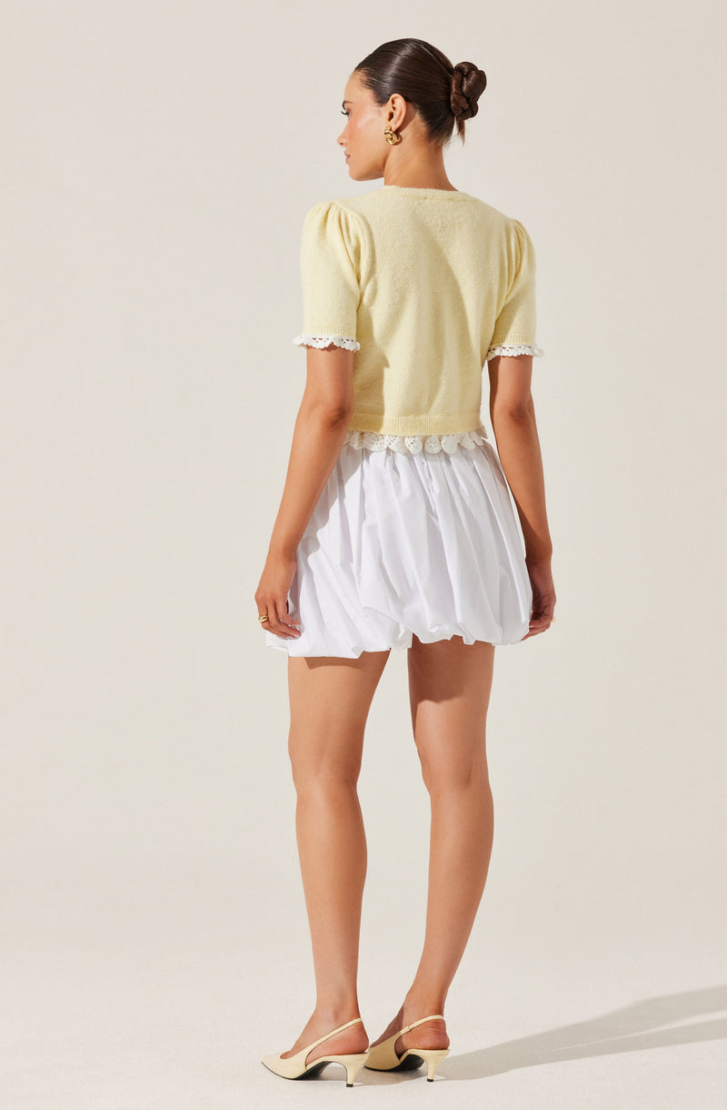 A woman is wearing a short-sleeved, light yellow sweater with puffed sleeves that have lace trim at the edges. The sweater has a ribbed hem. She is also wearing a white, loose, puffy skirt with a gathered, bubble-like silhouette. She has on beige, open-toe heels with ankle straps.