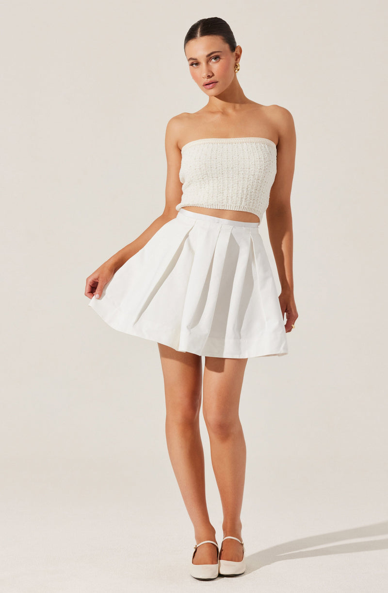 The woman is wearing a strapless white knitted crop top with a textured pattern and a high-waisted white pleated skirt that flares out above the knees. She is also wearing white closed-toe shoes with ankle straps and gold earrings.