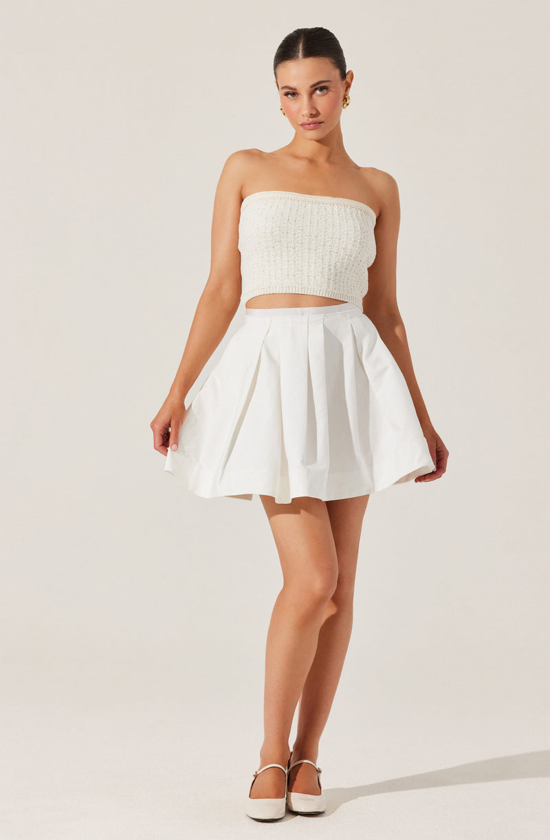 A woman is wearing a strapless, knitted, cream-colored crop top and a high-waisted, white pleated skirt with a tied waist. She is also wearing white high-heeled shoes.
