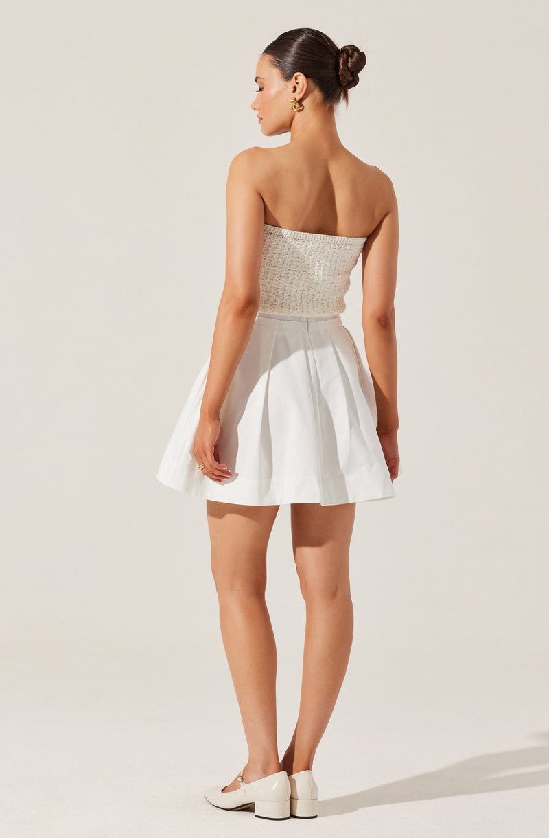 A woman is wearing a strapless, textured beige top paired with a high-waisted, white, pleated mini skirt. She has on white shoes with a chunky heel and gold earrings. Her hair is styled in a low, neat bun.