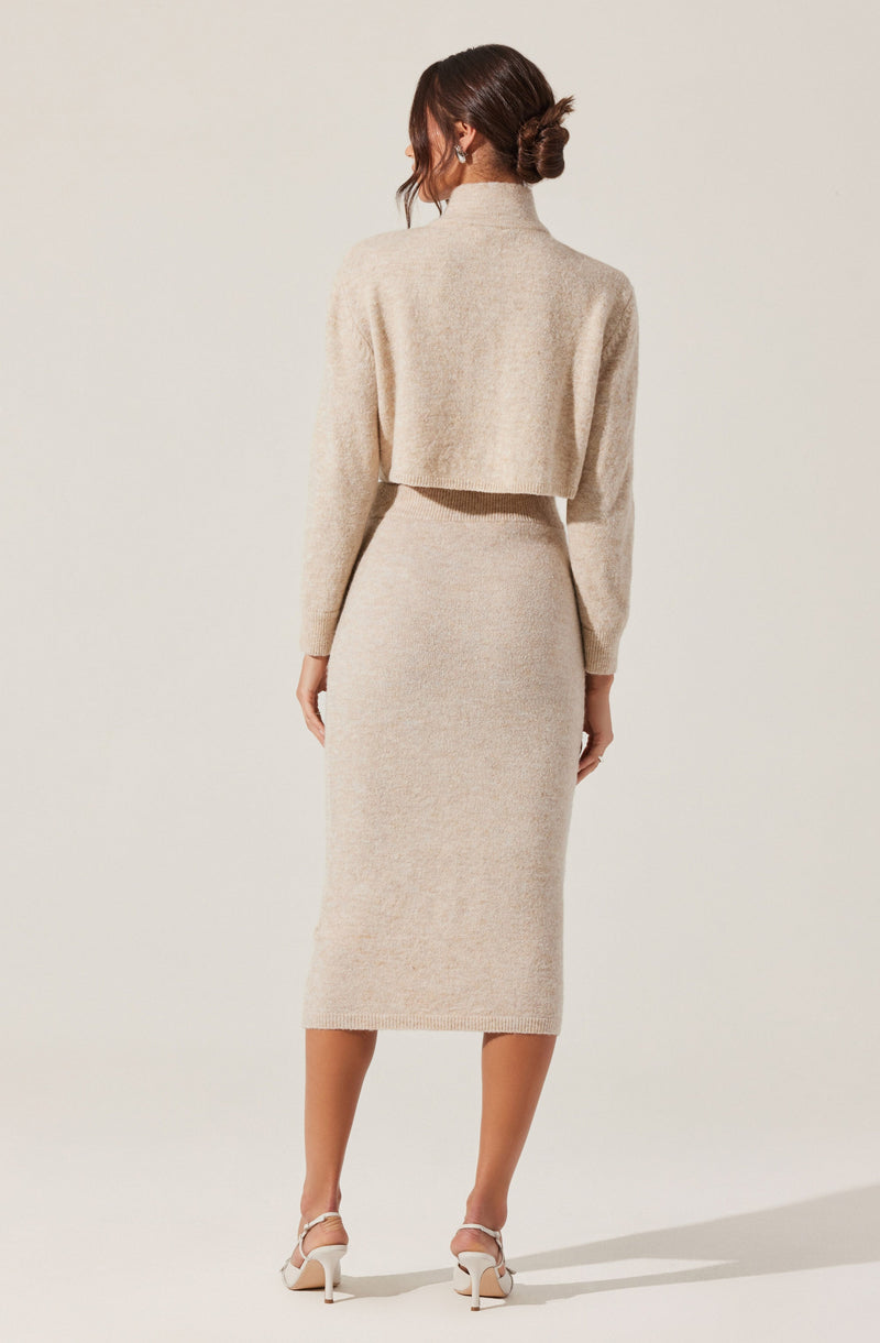 A woman is wearing a beige, knitted two-piece outfit consisting of a cropped turtleneck sweater with long sleeves and a matching high-waisted, midi-length skirt. She has her hair styled in a low bun and is wearing white high-heeled shoes.