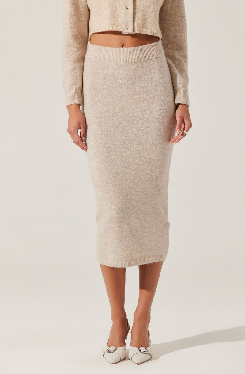 A woman is wearing a beige, high-waisted, knit midi skirt that reaches below her knees. She is also dressed in a matching beige, cropped cardigan with long sleeves and a button-up front. Her shoes are white, pointed-toe heels with a decorative bow on the front.