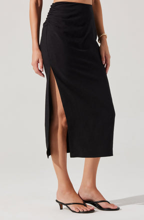 A woman is wearing a black midi skirt with a high slit on the left side, revealing her leg. The skirt appears to be made of a lightweight fabric with a slight Gathered detail on the side. She is also wearing black strappy sandals with a small kitten heel and a gold bracelet on her right wrist.