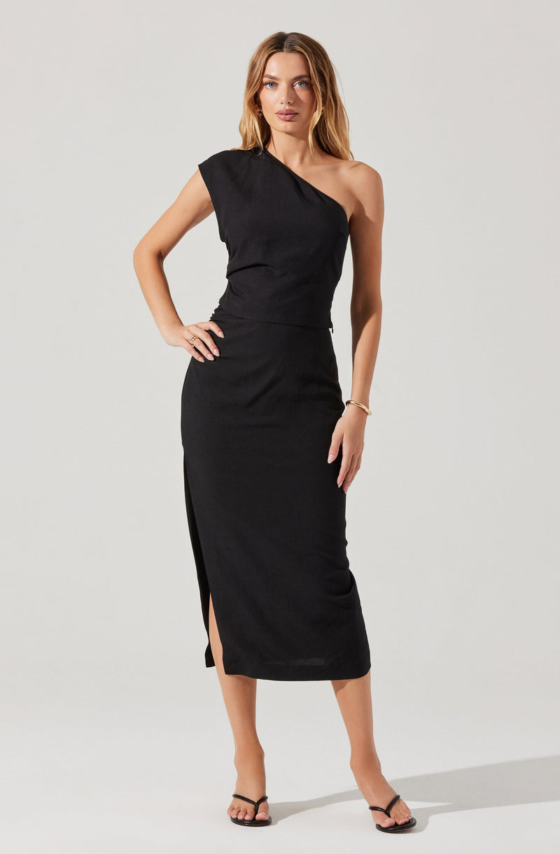 A woman is wearing a black dress with a one-shoulder design. The dress is fitted at the waist and extends down to mid-calf length, featuring a slit on the left side. She is also wearing black heeled sandals and a simple bracelet on her right wrist.