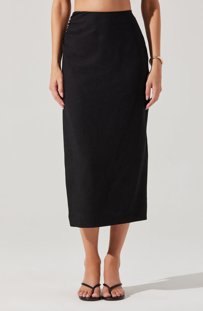 A woman is wearing a high-waisted, ankle-length black skirt made of a smooth, possibly satin-like fabric. She has paired it with black strappy sandals and a gold bracelet on her right wrist. The image focuses on her lower body from the waist down.