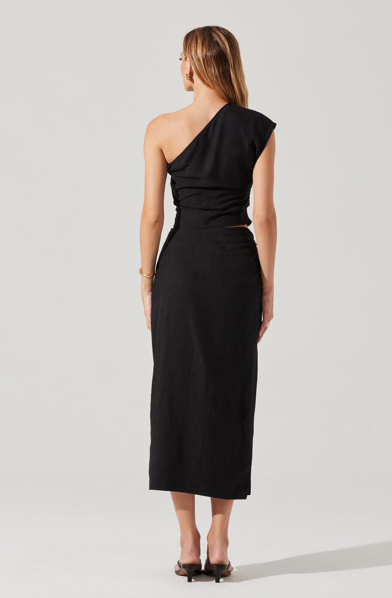A woman is wearing a black evening dress with an asymmetrical, one-shoulder top that drapes over one shoulder, leaving the other shoulder bare. The dress is fitted at the waist and extends to a long, straight, ankle-length skirt. She is accessorized with earrings, a bracelet, and open-toed black heels.
