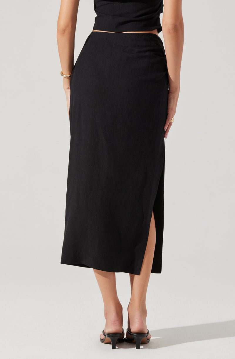 A woman is wearing a black, high-waisted, ankle-length skirt with a front slit on the left side. She pairs it with black open-toe heels and a black short-sleeved top. She accessorizes with a bracelet on her right wrist and a ring on her left hand.