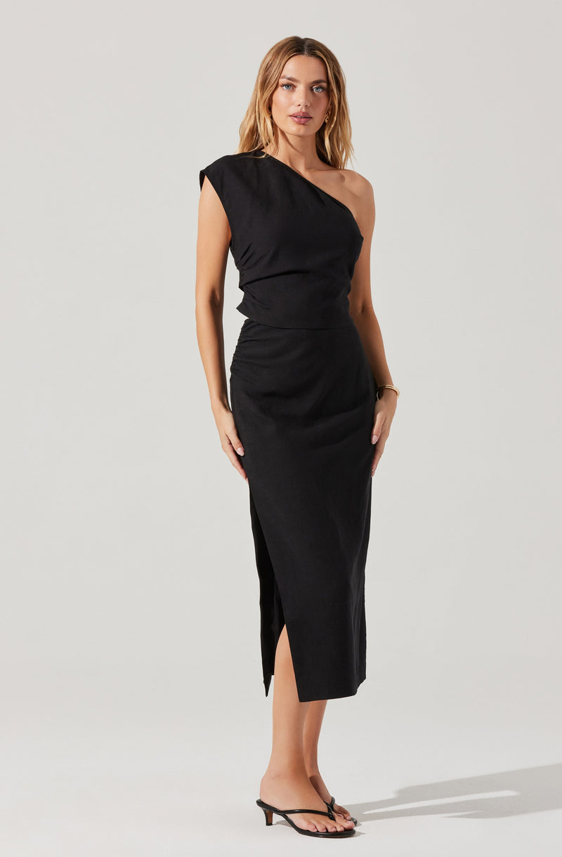 A woman is wearing a black dress with a one-shoulder design. The dress has a fitted bodice and a long, straight skirt that reaches mid-calf, with a small slit on the side. She is also wearing black, open-toed high-heeled sandals.