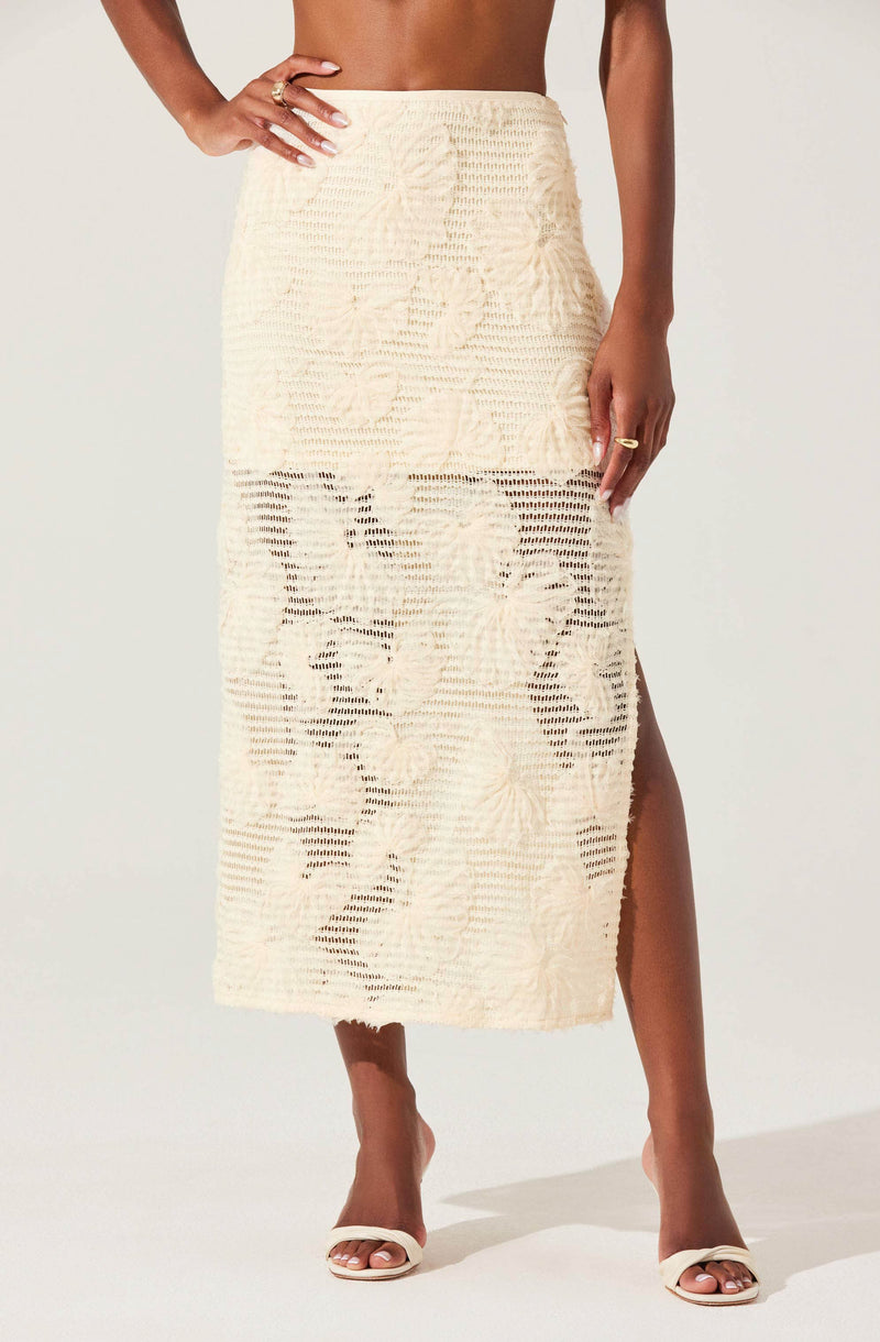 A woman is wearing a long, off-white crochet skirt featuring a floral and portrait-like design. The skirt has a side slit revealing her leg and she pairs it with open-toe heels. She is accessorizing with rings on her fingers.