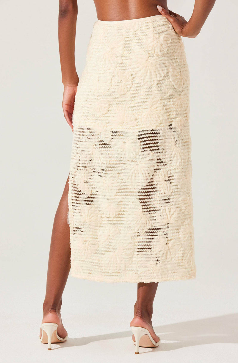 A woman is wearing a long, cream-colored skirt made of a textured, open-knit fabric. The skirt features large floral patterns knitted into the material and has a slit on one side that reaches above the ankle. She is also wearing nude high-heeled shoes.