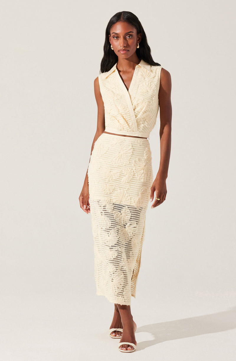 A woman is wearing a sleeveless, cream-colored, textured crop top with a V-neckline and structured shoulders. She pairs it with a matching high-waisted, ankle-length skirt made of the same textured fabric, which has a sheer, patterned overlay. She completes the outfit with open-toe heels.