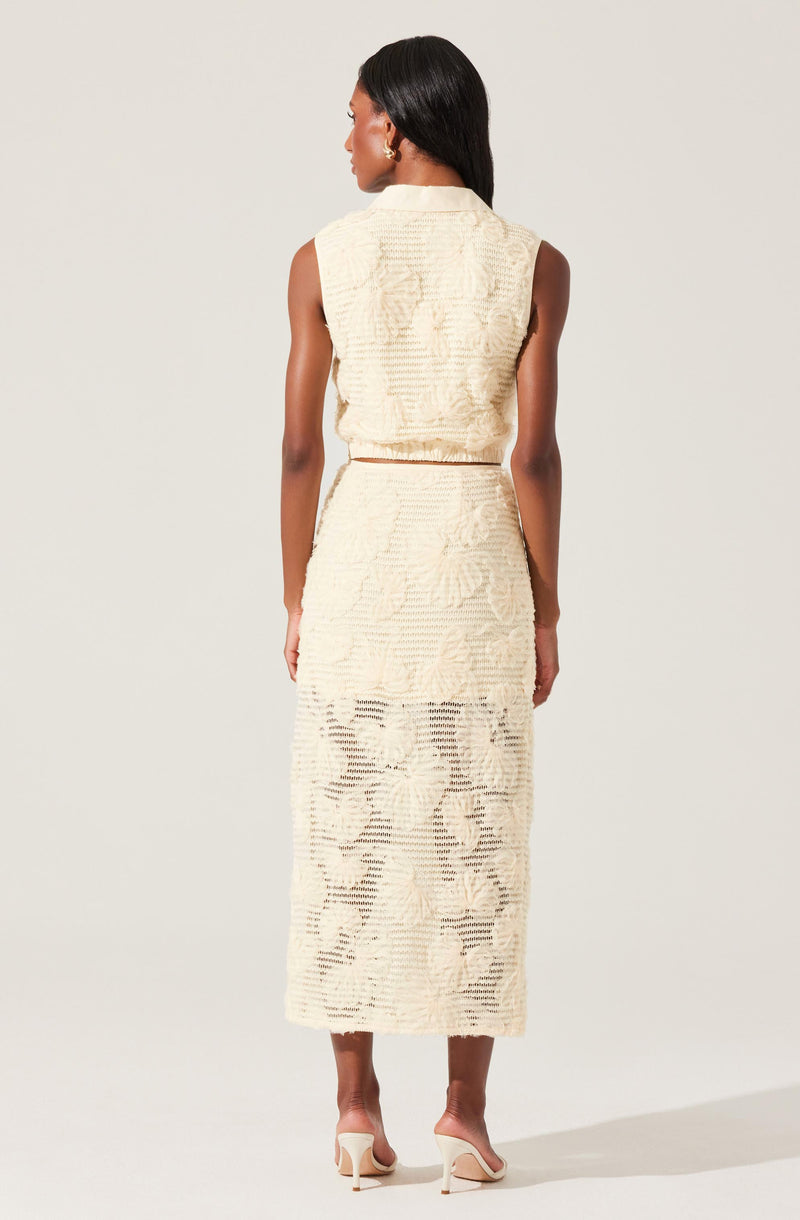 A woman is wearing a sleeveless, cream-colored textured two-piece outfit consisting of a cropped top with wide shoulders and a high-waisted, long skirt that extends to her ankles. The fabric has a subtle floral pattern with openwork details. She is also wearing nude high-heeled shoes.