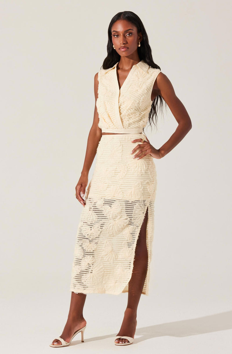 A woman is wearing a sleeveless, cream-colored two-piece outfit with textured fabric. The top has a V-neckline with a layered, wrap-style design. The matching skirt is high-waisted and midi-length, featuring a slit on one side. She is also wearing open-toed, nude high heels and small gold earrings. Her long, wavy hair is down, and she has a confident pose with one hand on her hip.