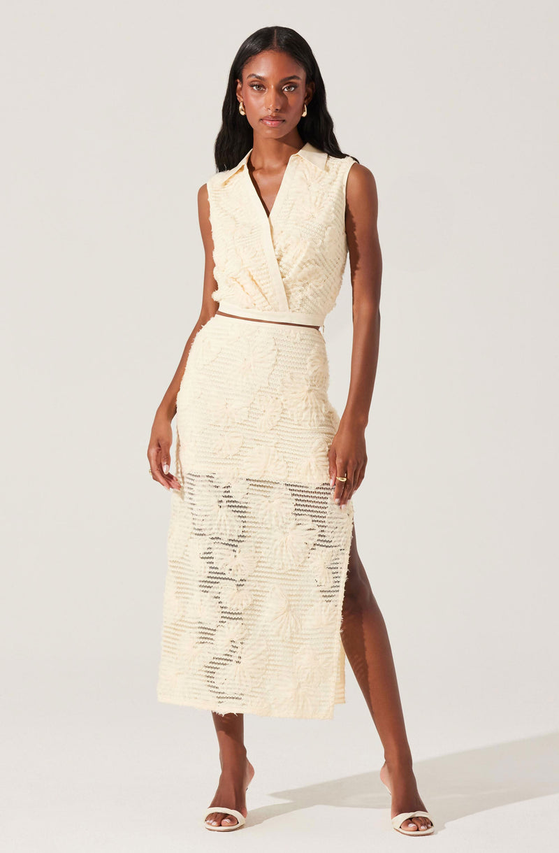 A woman is wearing a sleeveless, cream-colored set consisting of a textured wrap-style top with a deep V-neck and a long, matching skirt with a slit on one side. The fabric has a subtle, patterned texture. She is also wearing open-toe, high-heeled sandals.