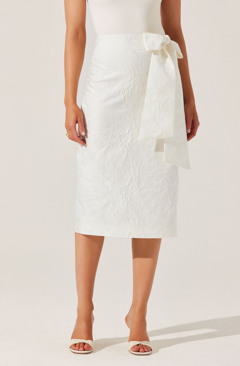 The image shows a woman wearing a white, textured midi skirt with a large, matching waist tie bow on the right side. She is also wearing a sleeveless, fitted white top and open-toed, high-heeled sandals in a neutral color.