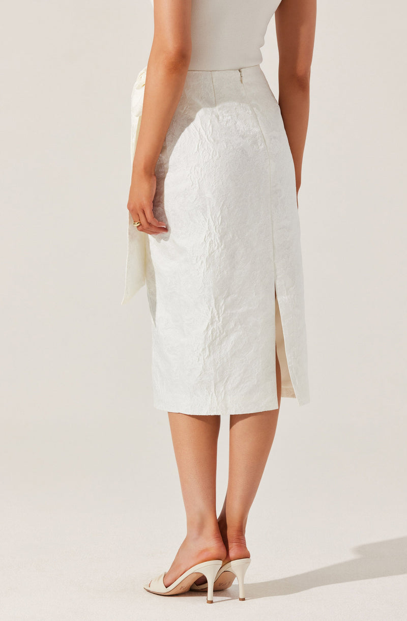 A woman is wearing a high-waisted, knee-length beige skirt with a front slit. The skirt appears to be made of a textured fabric. She is also wearing high-heeled sandals in a light color. Her top is sleeveless and a neutral color, matching the minimalist and elegant style of the outfit.