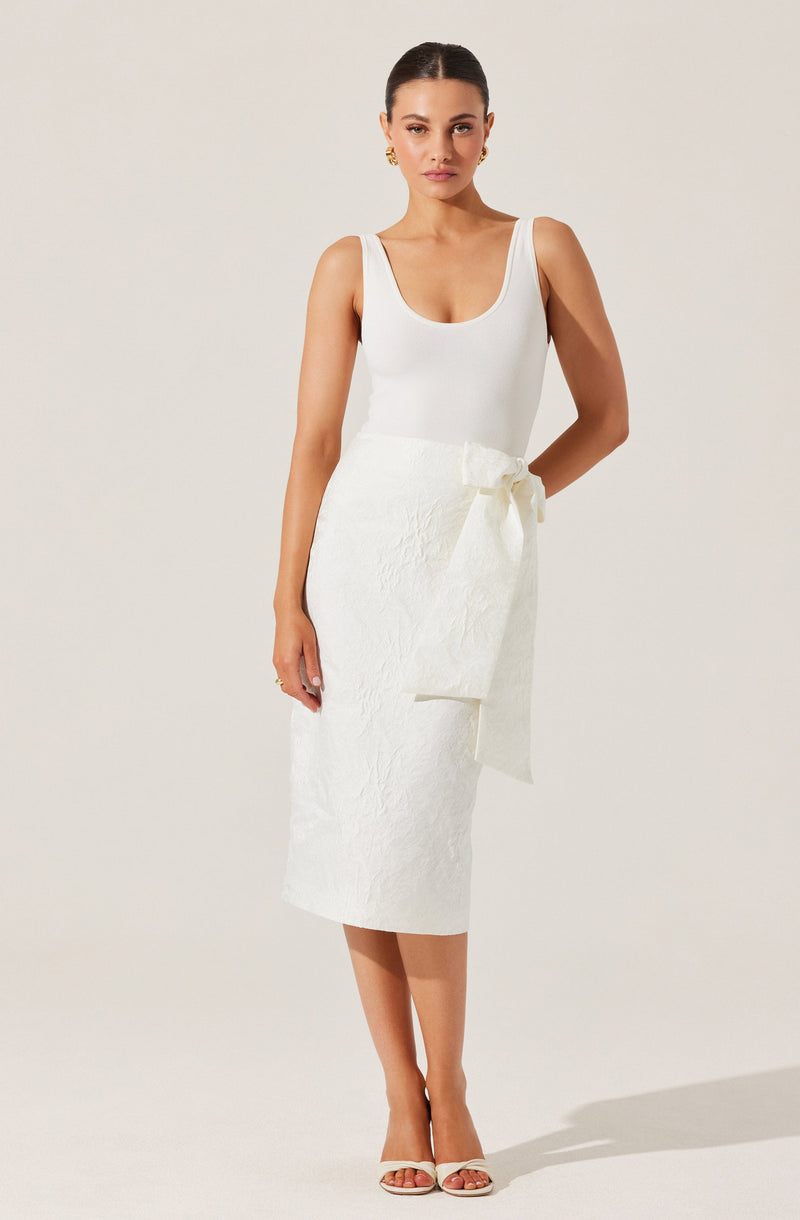 A woman is wearing a sleeveless white top with wide shoulder straps and a scoop neckline, paired with a high-waisted, mid-length white skirt featuring a textured fabric and a large bow tied at the waist. She is also wearing white open-toe high heels and gold earrings.