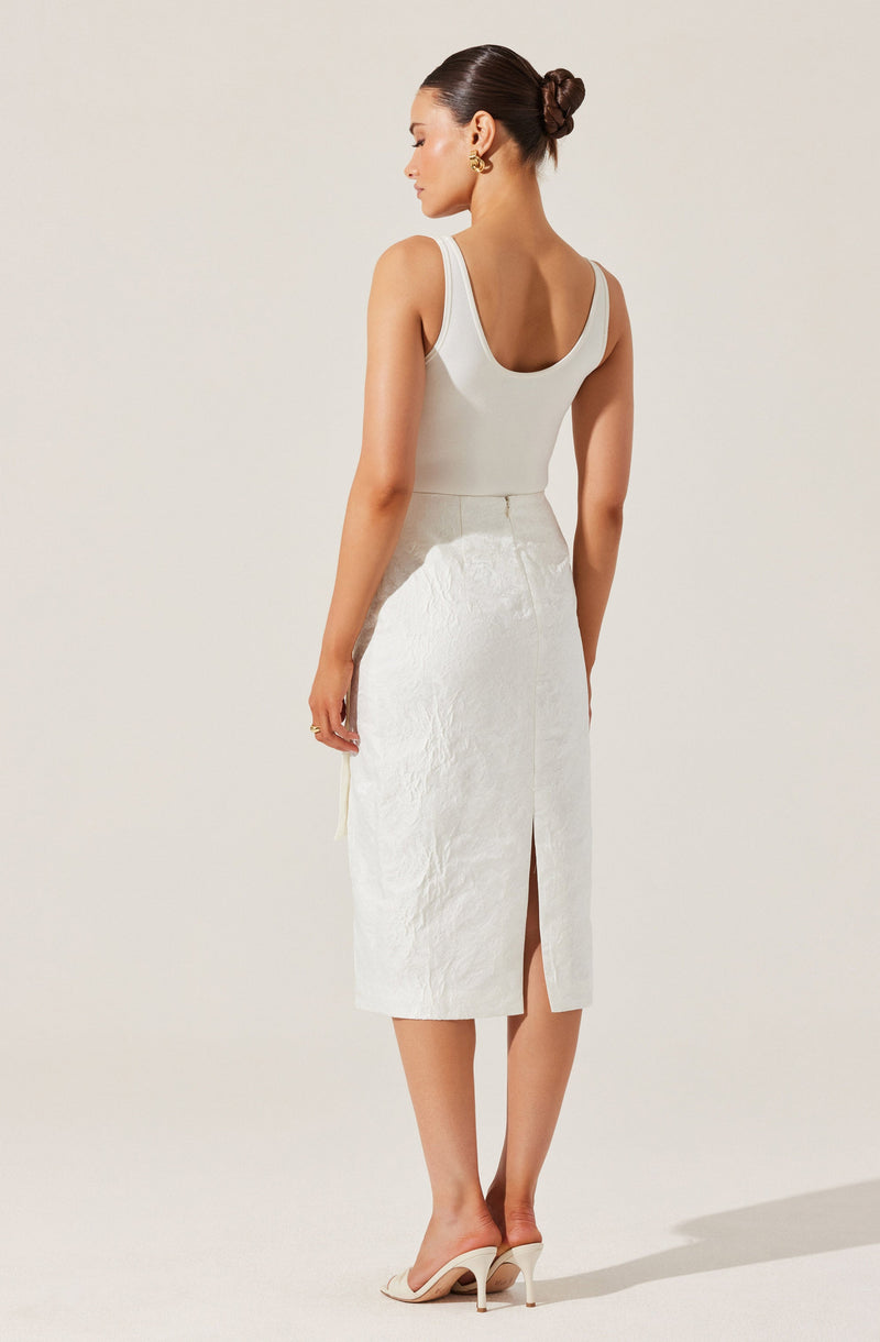 A woman is wearing a sleeveless, fitted, white top with a scoop neckline, paired with a high-waisted, mid-length white skirt that has a front slit. She is also wearing light-colored high-heeled shoes and gold hoop earrings. Her hair is styled in a polished, low bun.