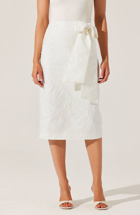 A woman is wearing a white, textured, midi-length skirt with a large side bow tied at the waist. She is also wearing white open-toe high heels.