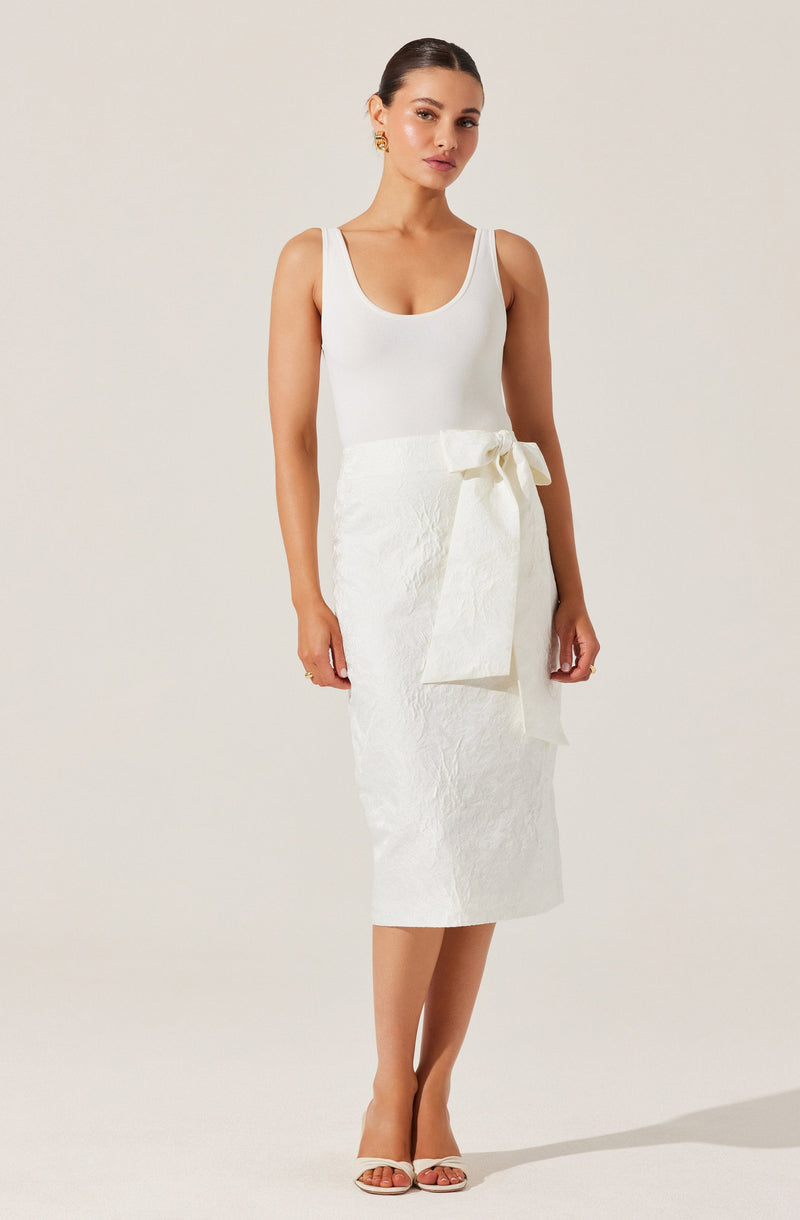 A woman is wearing a sleeveless white top with a scoop neckline paired with a high-waisted, textured white skirt that has a large bow tied at the waist. She is also wearing white open-toe high-heeled sandals.