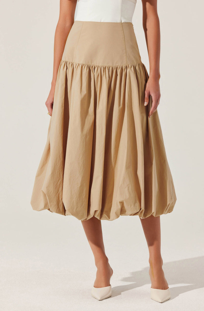A woman is wearing a high-waisted beige midi skirt with a gathered, voluminous bottom section that creates a bubble effect. The skirt appears to be made of a lightweight, slightly shiny fabric. She is also wearing a white top tucked into the skirt and white pointed-toe shoes.