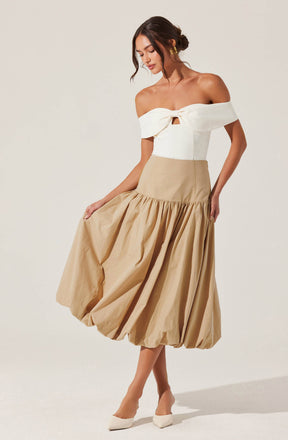 A woman is wearing an off-the-shoulder white top with a front tie detail and a cut-out at the center of the chest. She pairs it with a high-waisted, beige, full-volume midi skirt with a gathered, billowy silhouette. She completes her outfit with white pointed-toe heels.