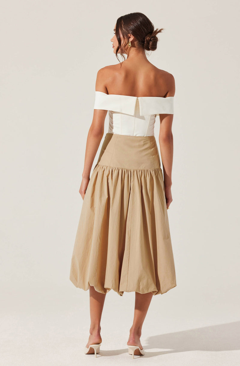 The woman is wearing an off-the-shoulder white top with a wide band that drapes over her upper arms and features a back zipper closure. She pairs it with a high-waisted beige skirt that has a fitted waistband and a gathered, voluminous, midi-length hem.