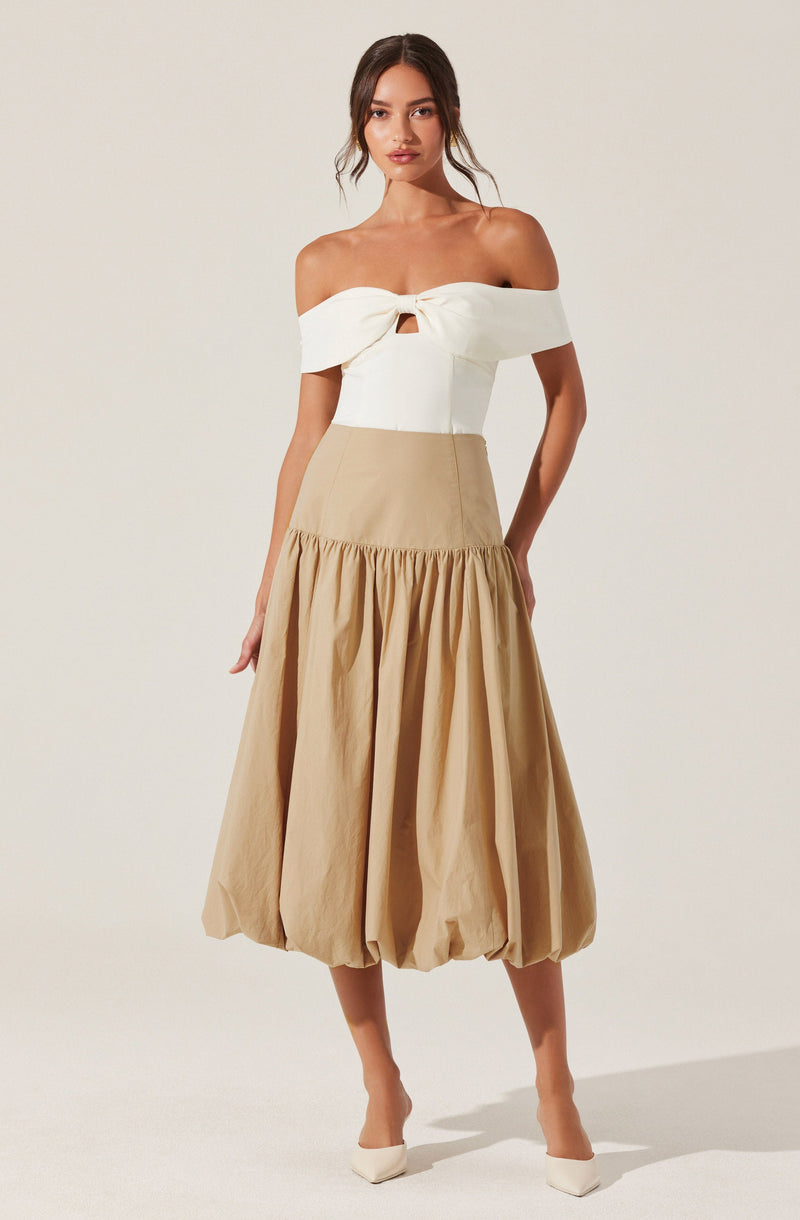 A woman is wearing an off-the-shoulder white top with a front knot and a small cutout below the knot. She pairs it with a high-waisted, beige, balloon-style skirt that has a gathered, voluminous design at the hem. She completes the outfit with white pointed-toe shoes.
