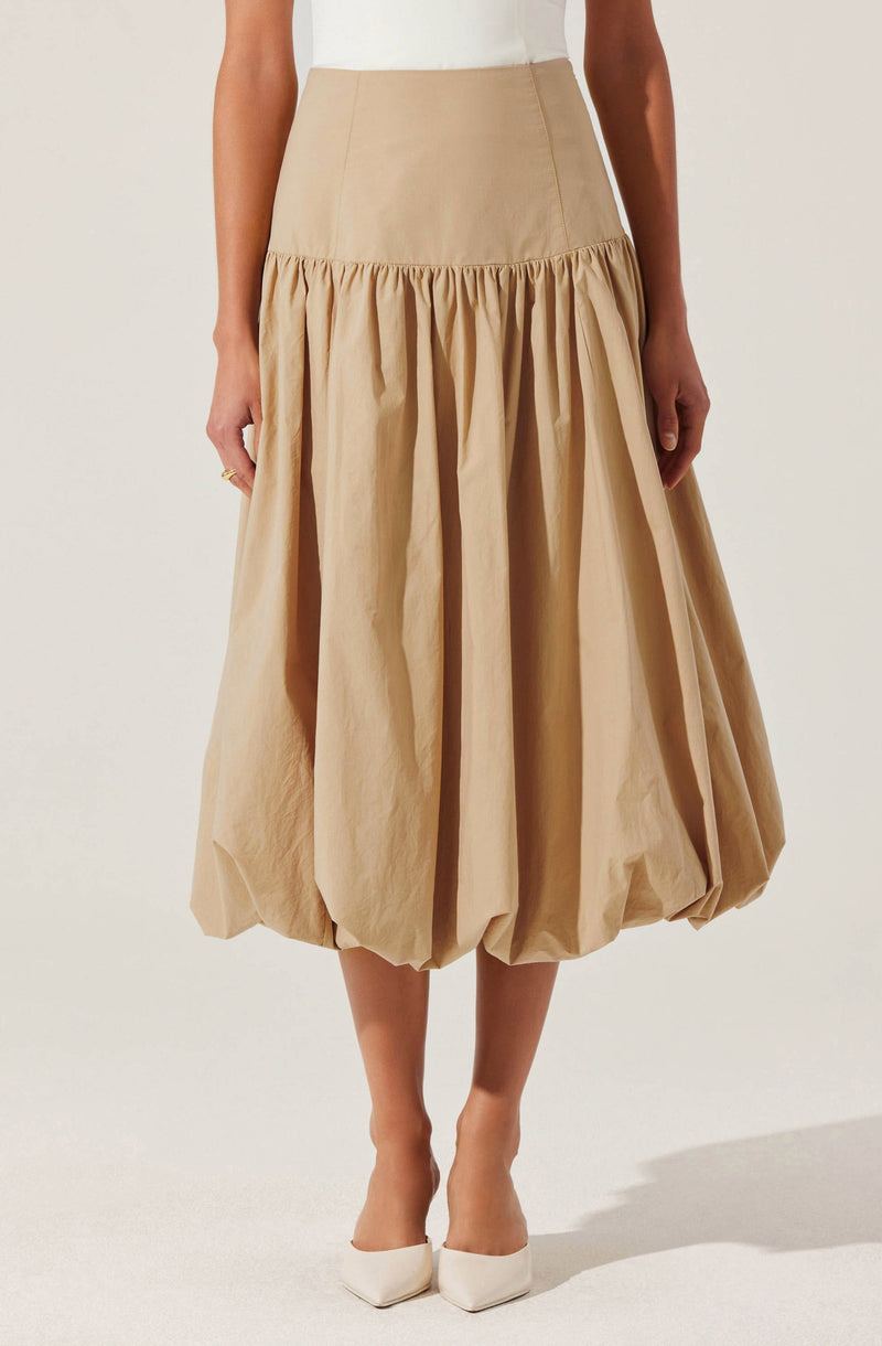 A beige high-waisted skirt with a gathered, voluminous, balloon-style silhouette that falls to mid-calf length. The skirt features a fitted waistband and gathered fabric creating pleats and a bubbly effect at the hem. The wearer pairs it with white pointed-toe heels.