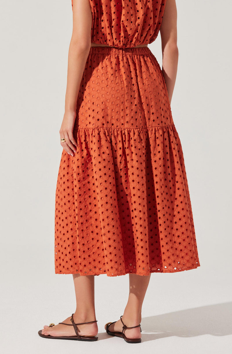 A woman is wearing an orange, sleeveless, midi-length dress with a drawstring waist. The dress features a textured pattern of evenly spaced black polka dots throughout the fabric. The skirt portion of the dress has a gathered, tiered design with a slight flare at the hem. She is also wearing flat sandals with ankle straps.