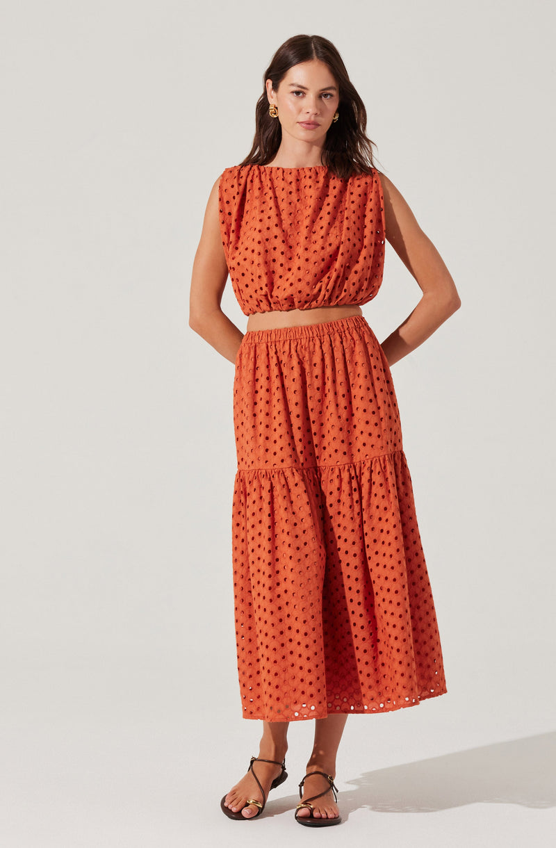 A woman is wearing a sleeveless, orange two-piece outfit made of a fabric with a black polka dot pattern. The top is a loose, cropped blouse with a gathered neckline and armholes. The skirt is high-waisted, flowy, and midi-length, featuring a tiered design with gathered fabric at the seams.