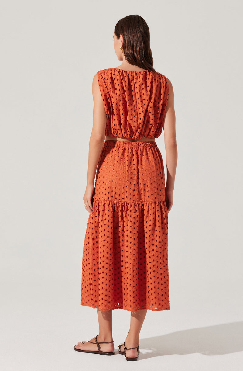 The woman is wearing a sleeveless, rust-colored dress with a textured, dotted pattern. The dress features a gathered neckline and a high elastic waistband, with a tiered, flowing skirt that reaches mid-calf length.