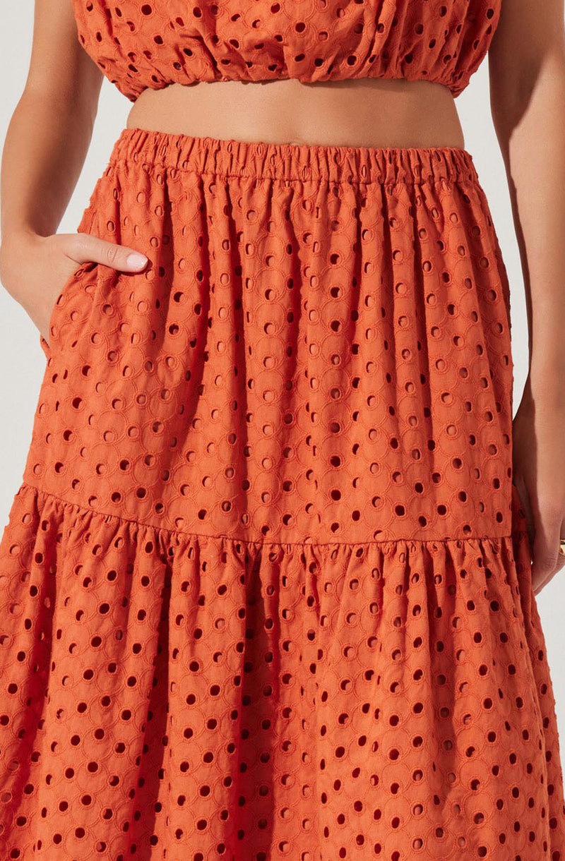 A woman is wearing a matching two-piece set made of orange fabric with a pattern of evenly spaced, circular cutouts. The top is a sleeveless crop with an elasticized band at the hem, and the skirt is a high-waisted, tiered design with an elastic waistband and gathered fabric at the tiers. The skirt has large side pockets, and the woman's hand is resting in the pocket on her right side.