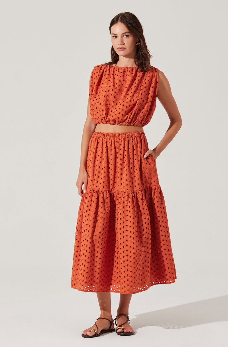A woman is wearing a two-piece outfit made of orange fabric with a black polka dot pattern. The top is a sleeveless, loose-fitting blouse with a round neckline and gathered fabric at the shoulders, creating a slightly puffed effect. The skirt is a long, tiered midi length with an elastic waistband, and it features a ruffled tier near the hem. She is also wearing black strappy sandals.