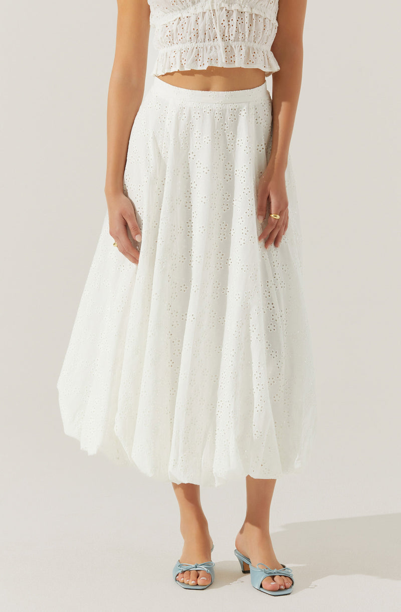 A woman is wearing a white, eyelet, ankle-length skirt with a full, gathered silhouette. She pairs it with a matching white, sleeveless, eyelet top that has a ruffled, cropped design with a smocked waist. She is also wearing pale blue, open-toe sandals with a bow embellishment on the front.