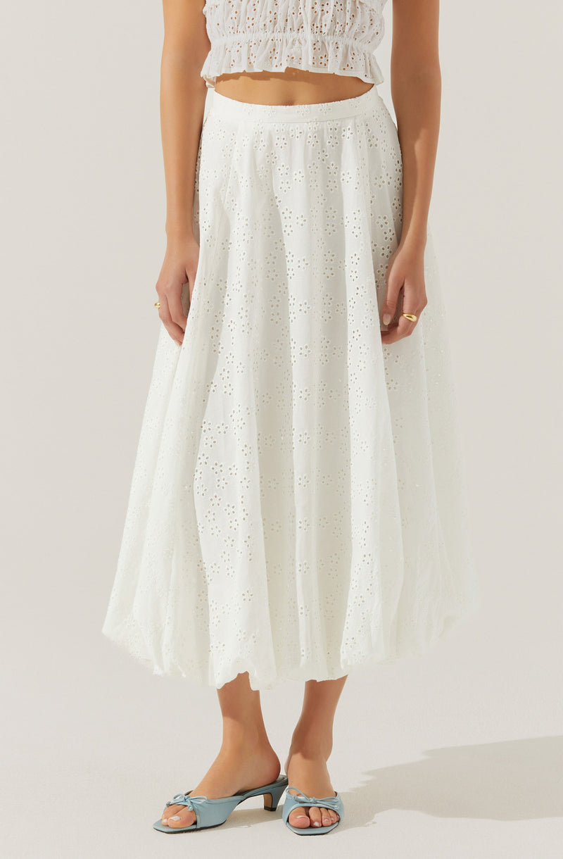 A woman is wearing a white, eyelet, ankle-length skirt with a flowy fit. She pairs it with a matching white eyelet crop top with a ruffled hem. She is also wearing light blue open-toe sandals with a small heel and a bow detail on the front.