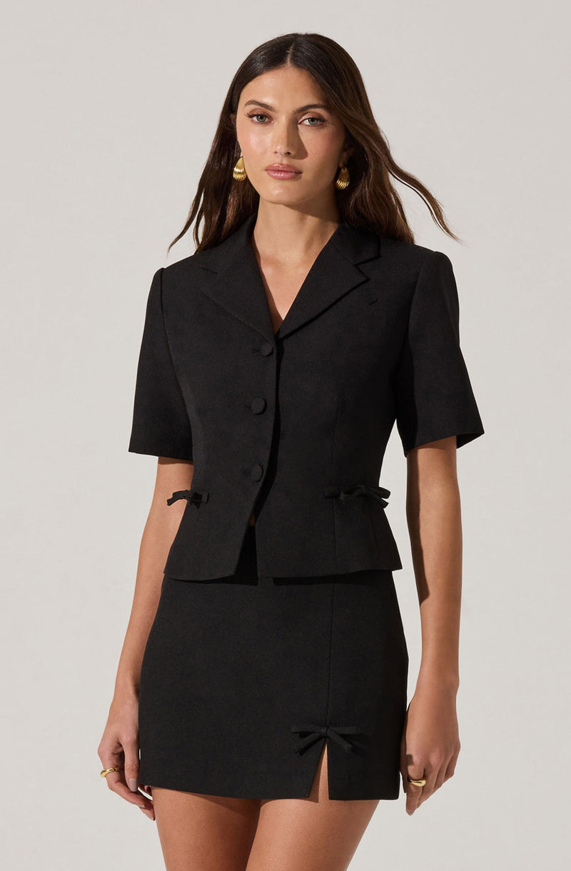 A woman is wearing a tailored black short-sleeve blazer with a notched lapel collar, three buttons down the front, and small bows at the waist on each side. She pairs it with a matching black mini skirt that features a front slit and a decorative bow near the hem. She has gold hoop earrings and rings on her fingers.