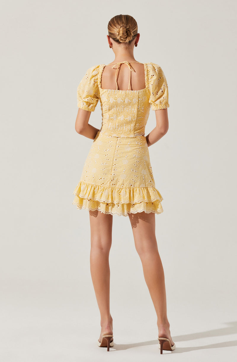 A woman is wearing a yellow dress with puffed short sleeves, a square neckline, and a fitted bodice. The dress features eyelet embroidery throughout, a ruffled hemline, and buttons running down the back. She is also wearing nude high-heeled shoes and has her hair styled in a neat bun.