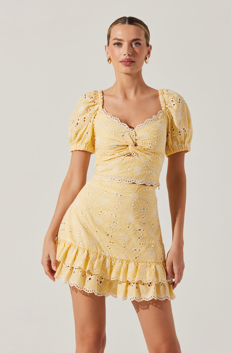 The woman is wearing a light yellow, eyelet embroidered two-piece outfit. The top has short, puffed sleeves with gathered cuffs, a square neckline with scalloped edges, and a front tie detail. The matching skirt is high-waisted, fitted through the hips, and features a ruffled hem with eyelet embroidery matching the top.