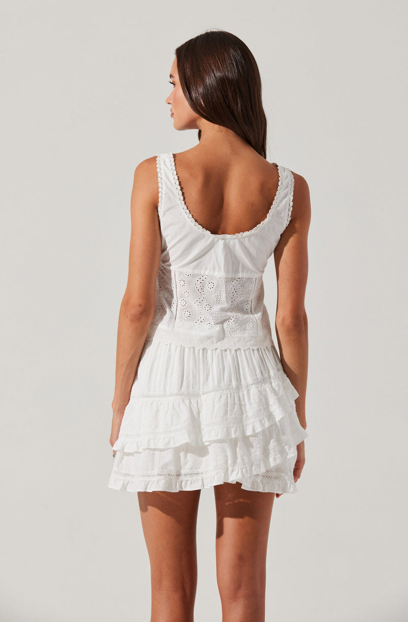 A woman is wearing a sleeveless, white dress with a scoop neckline and lace trim on the shoulder straps. The dress features eyelet embroidery on the upper back panel and has multiple layered ruffles on the skirt, creating a tiered effect.