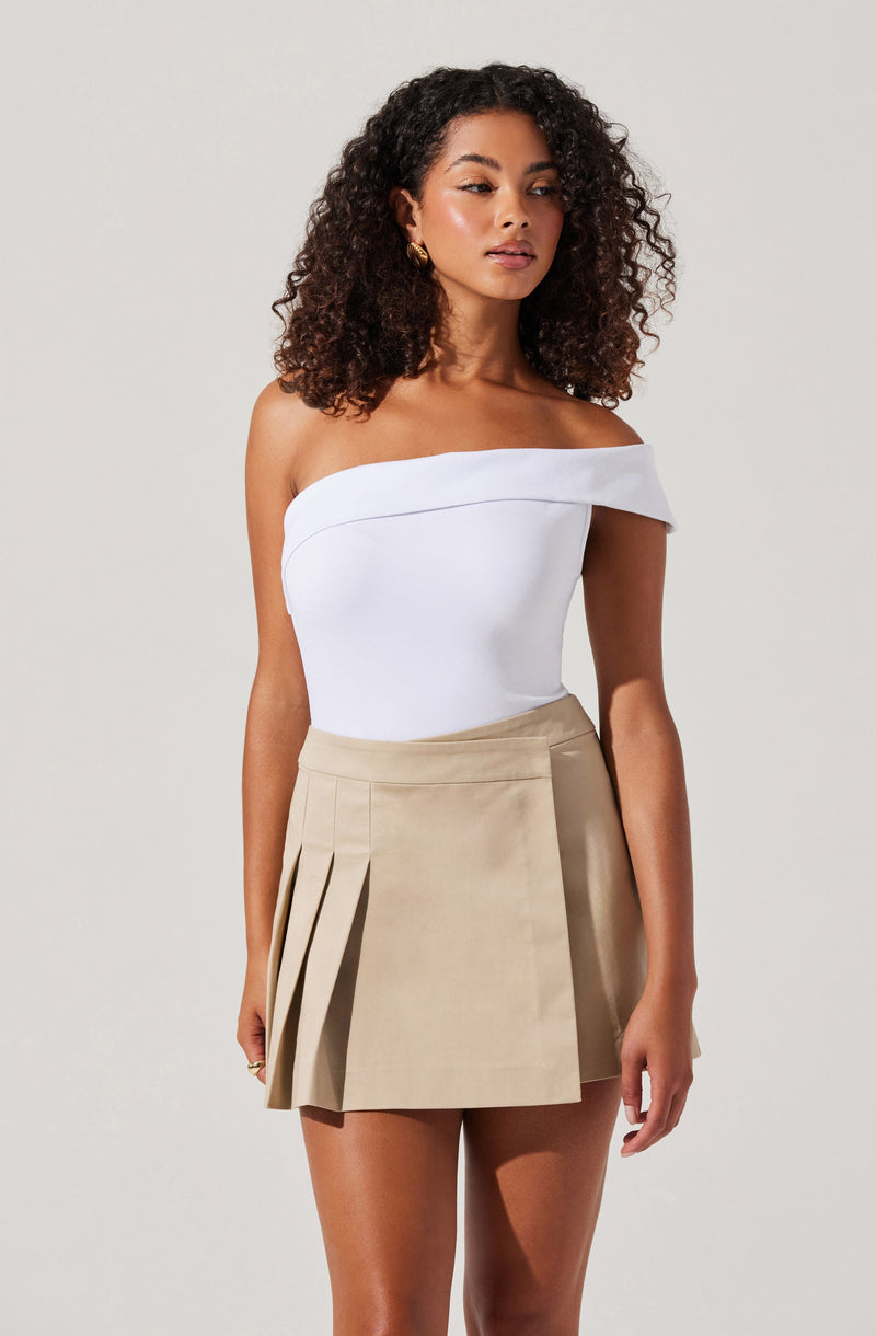 A woman is wearing a white, off-the-shoulder, fitted top with a wide neckline that drapes over her shoulders. She has paired it with a beige pleated mini skirt that features multiple pleats on the left side, creating a layered look. The outfit is complemented by gold earrings.