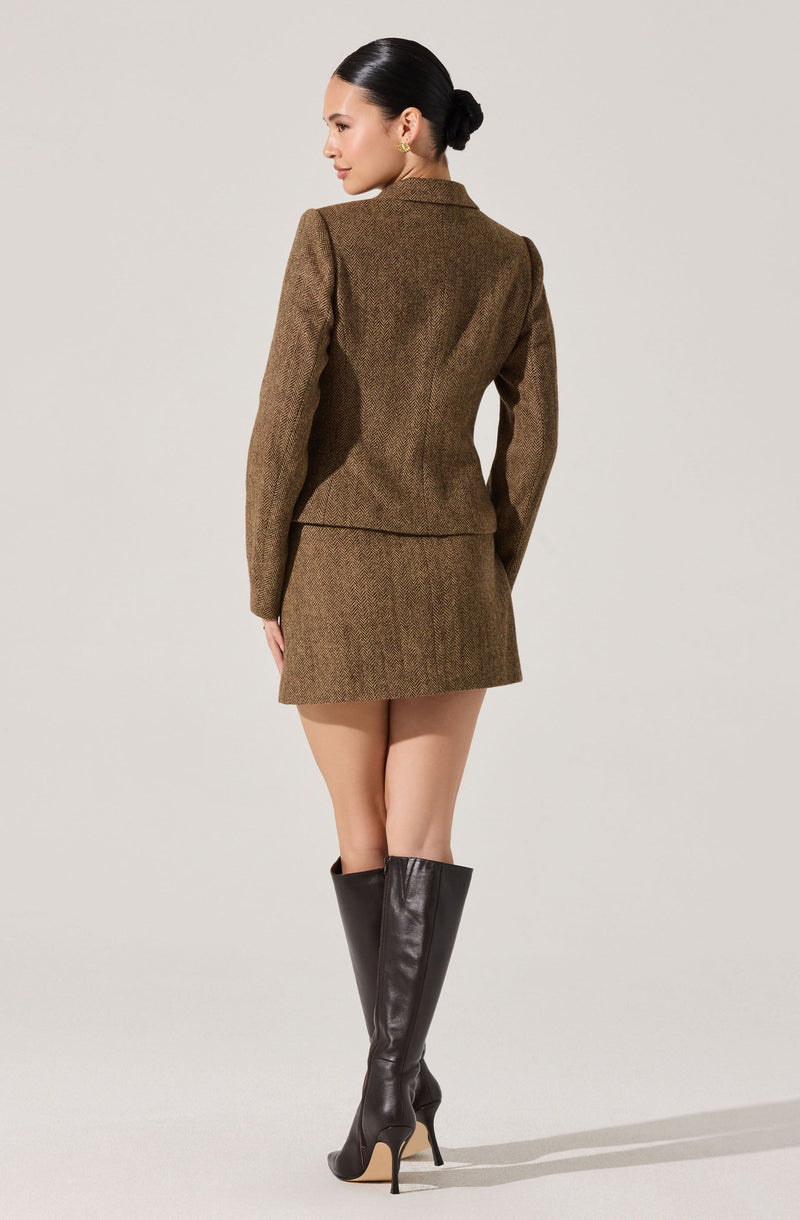 A woman is wearing a tailored, brown, textured blazer and a matching brown, textured mini skirt. She has her hair styled in a sleek bun and is accessorized with gold hoop earrings. She is also wearing tall, black, shiny leather high-heeled boots.